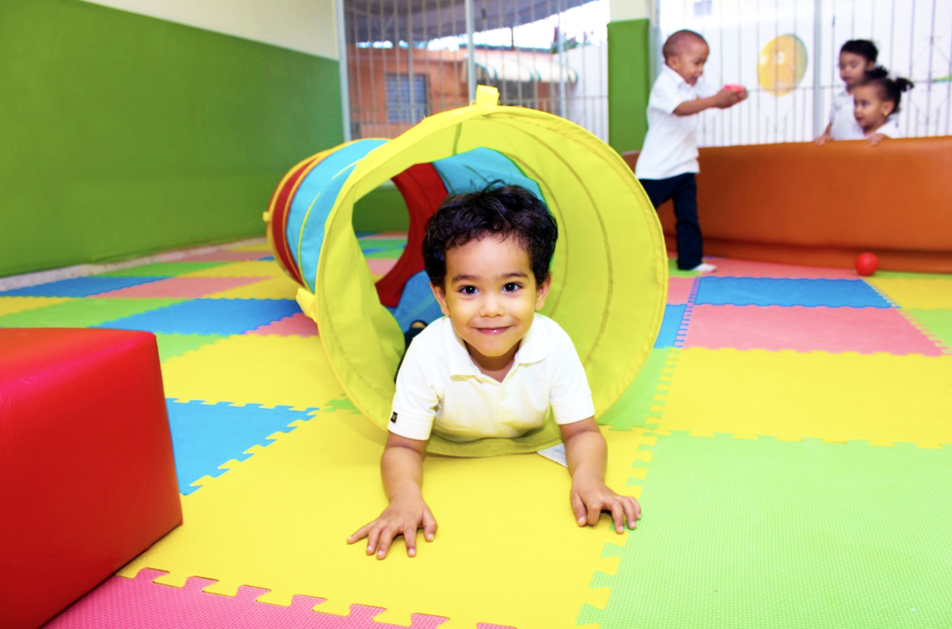 A young child crawls through a tube as other children play in the background indoors.