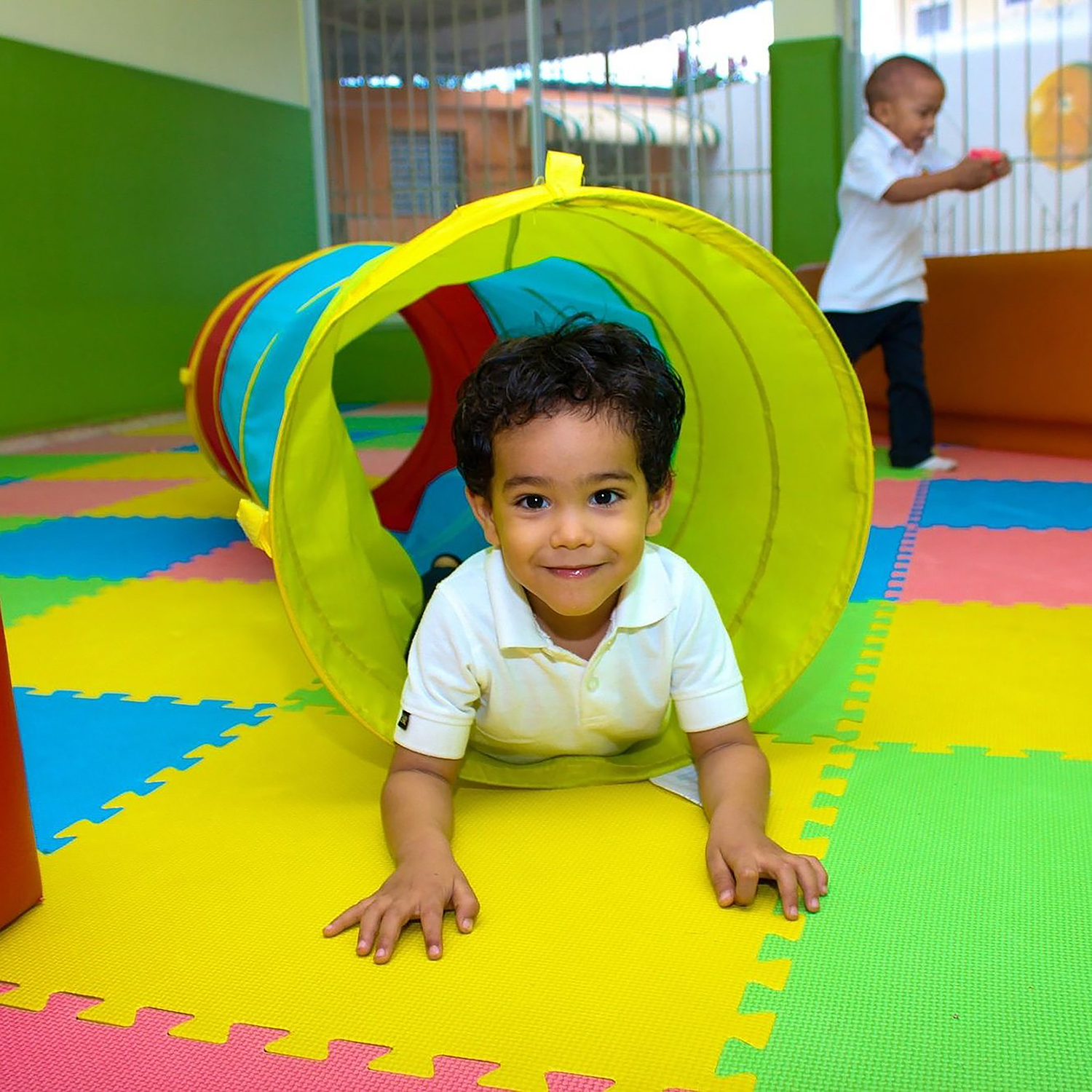 A young child crawls through a tube as other children play in the background indoors.